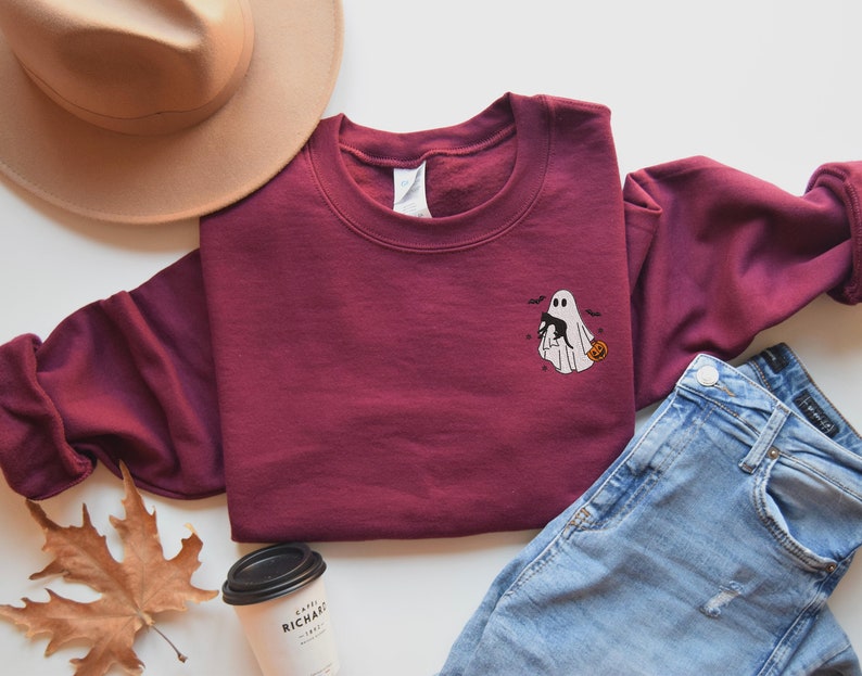 Op de afbeelding: Een bordeauxrood sweatshirt met een geborduurd ontwerp van een geest die een zwarte kat en een pompoen vasthoudt. Het ontwerp bevindt zich op de voorkant van het sweatshirt.