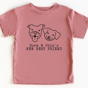 A pink t-shirt with a graphic design featuring two dog faces and the text "Sora & Odin's NEW BEST FRIEND".