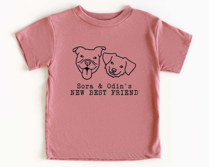 A pink t-shirt with a graphic design featuring two dog faces and the text "Sora & Odin's NEW BEST FRIEND".
