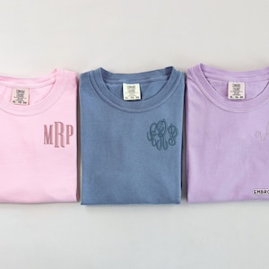 Puede incluir: Tres camisetas de manga corta en rosa claro, azul y lavanda. Cada camiseta tiene una etiqueta Comfort Colors en el cuello y un monograma bordado en el pecho izquierdo. La palabra EMBROIDERED aparece en un recuadro abajo a la derecha.
