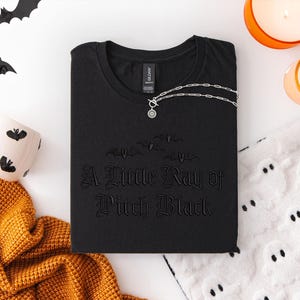 May include: Black t-shirt with the phrase "A Little Ray of Pitch Black" and bat graphics. A silver necklace with a circular pendant rests on the shirt. The image includes Halloween-themed decor, such as bat cutouts, candles, and a mug.