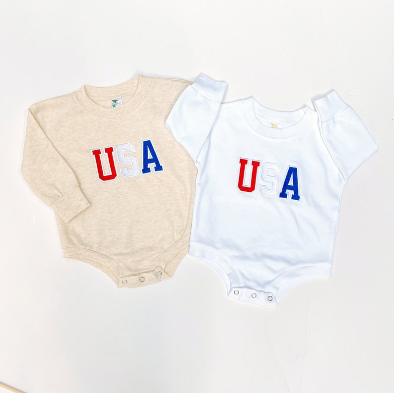 May include: Two long-sleeved baby onesies with the word "USA" in red, white, and blue embroidered on the front. One onesie is beige and the other is white.