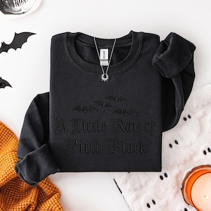 May include: Black crewneck sweatshirt with the phrase "A Little Ray of Pitch Black" in gothic lettering. The sweatshirt features a bat graphic above the text. A silver necklace with a sun pendant is visible. Halloween-themed apparel.