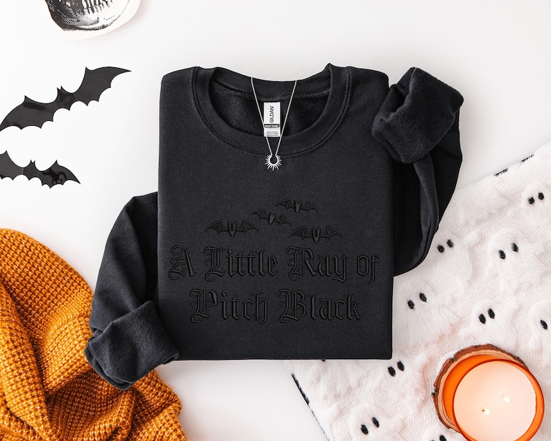 May include: Black crewneck sweatshirt with the phrase "A Little Ray of Pitch Black" in gothic lettering. The sweatshirt features a bat graphic above the text. A silver necklace with a sun pendant is visible. Halloween-themed apparel.