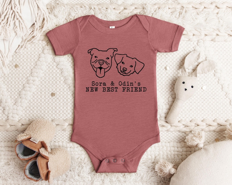 A pink baby onesie with a graphic of two dogs and the text "Sora & Odin's NEW BEST FRIEND" printed on it, along with a stuffed rabbit and a pair of shoes.