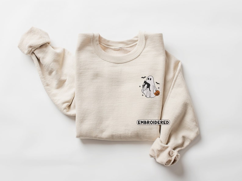 Op de afbeelding: Een beige sweatshirt met een geborduurd ontwerp van een geest die een zwarte kat en een pompoen vasthoudt. De tekst "EMBROIDERED" staat onder het ontwerp.