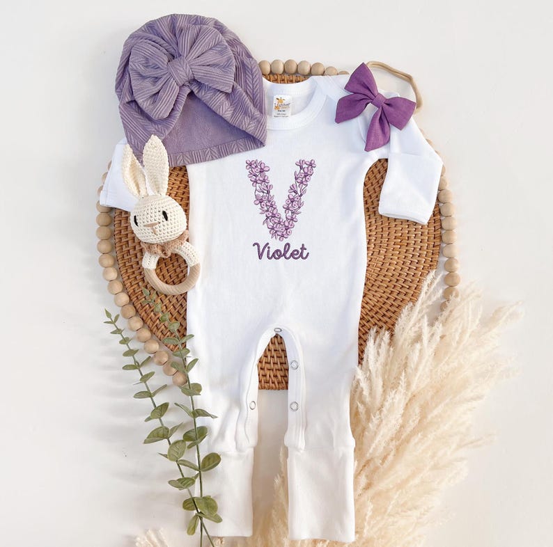 May include: White baby onesie with a purple floral letter V and the name "Violet" embroidered on the front. The onesie is paired with a purple turban and a purple bow headband.