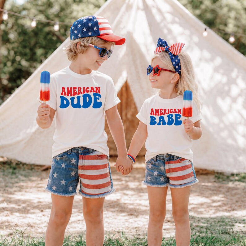 May include: Two children wearing matching outfits with patriotic themes. One child wears a white t-shirt with "AMERICAN DUDE" in blue, star-spangled shorts, and a baseball cap. The other wears a white t-shirt with "AMERICAN BABE" in blue, star-spangled shorts, and a headband.