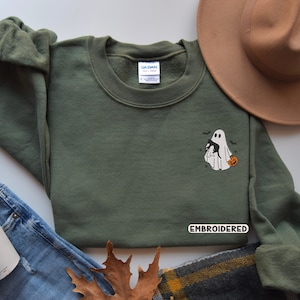 Op de afbeelding: Een groen sweatshirt met een geborduurd ontwerp van een geest die een zwarte kat en een pompoen vasthoudt. De tekst "EMBROIDERED" is zichtbaar op het sweatshirt.