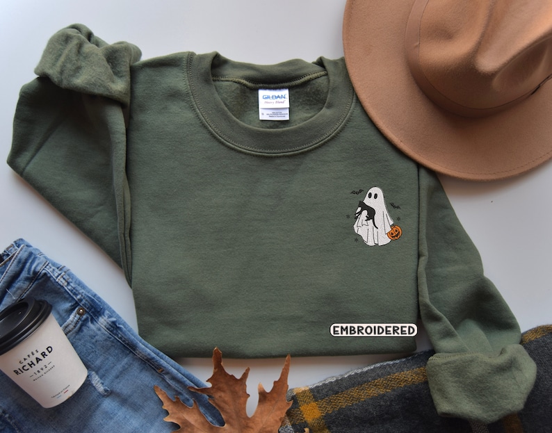 Op de afbeelding: Een groen sweatshirt met een geborduurd ontwerp van een geest die een zwarte kat en een pompoen vasthoudt. De tekst "EMBROIDERED" is zichtbaar op het sweatshirt.