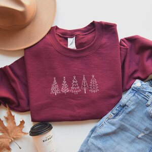 May include: A maroon sweatshirt with a white embroidered design of five stylised Christmas trees. The sweatshirt is laid flat on a white surface with a brown hat, a pair of blue jeans, and a coffee cup.