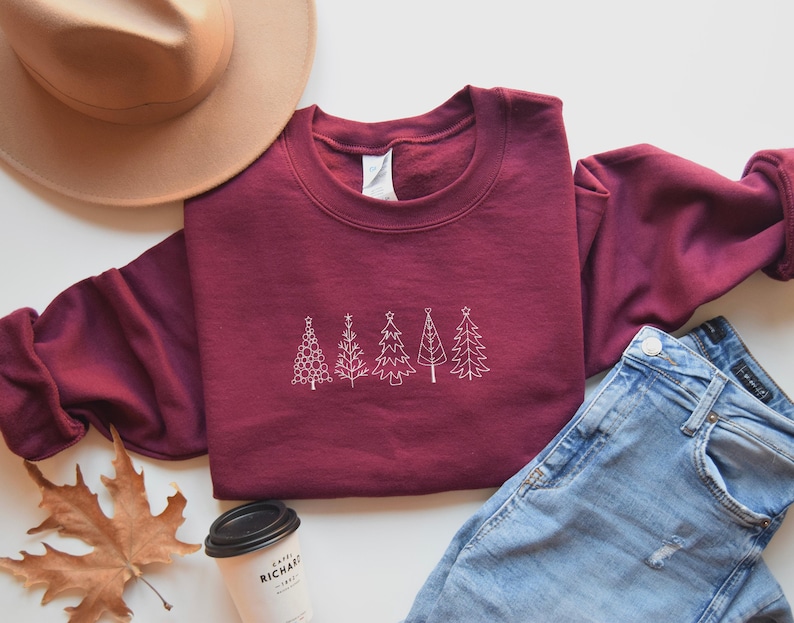 May include: A maroon sweatshirt with a white embroidered design of five stylised Christmas trees. The sweatshirt is laid flat on a white surface with a brown hat, a pair of blue jeans, and a coffee cup.