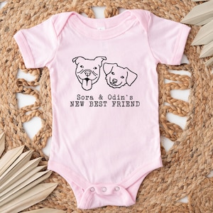 A pink baby onesie with a graphic of two dogs and the text "Sora & Odin's NEW BEST FRIEND" printed on it, placed on a woven mat with leaves.