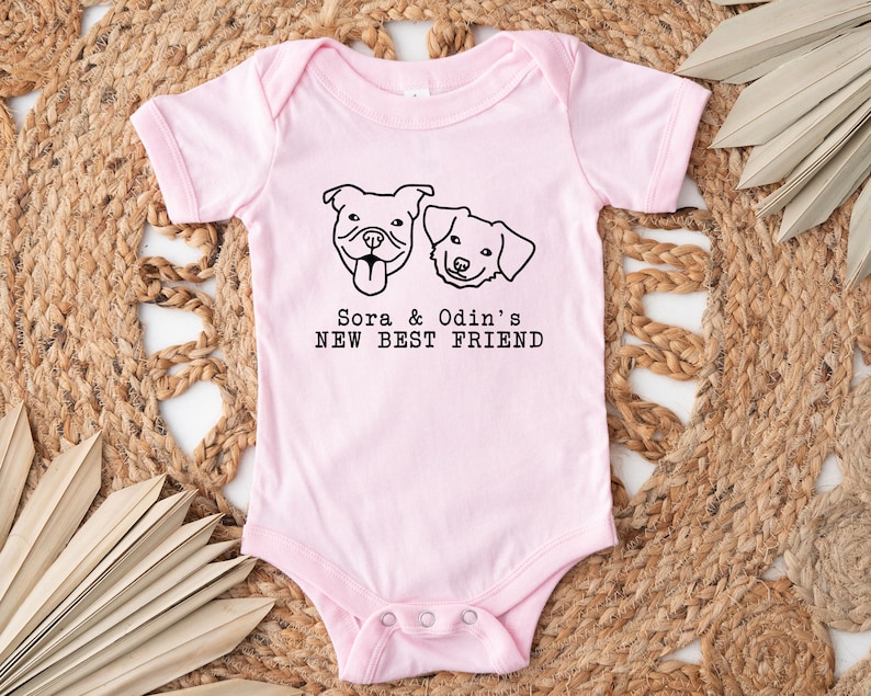 A pink baby onesie with a graphic of two dogs and the text "Sora & Odin's NEW BEST FRIEND" printed on it, placed on a woven mat with leaves.