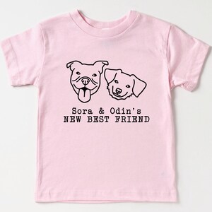 A pink t-shirt with a graphic design of two dog faces and the text "Sora & Odin's NEW BEST FRIEND" printed on it.