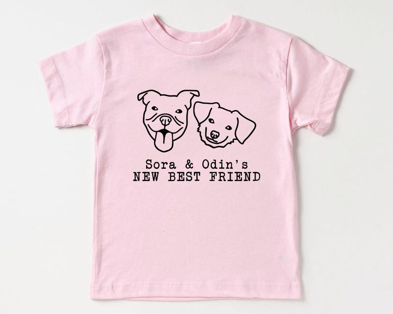 A pink t-shirt with a graphic design of two dog faces and the text "Sora & Odin's NEW BEST FRIEND" printed on it.