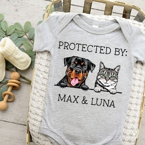 以下が含まれることがあります： 「PROTECTED BY: MAX & LUNA」というテキストと、ロットワイラーと猫のイラストが描かれたグレーのベビーロンパースです。
