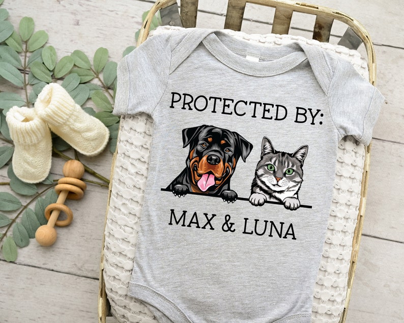 以下が含まれることがあります： 「PROTECTED BY: MAX & LUNA」というテキストと、ロットワイラーと猫のイラストが描かれたグレーのベビーロンパースです。
