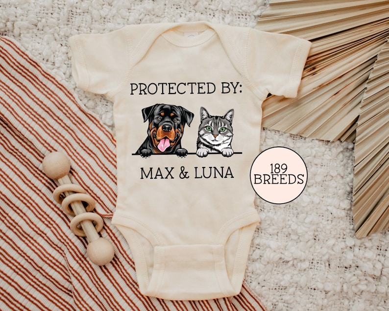 以下が含まれることがあります： 「PROTECTED BY: MAX & LUNA」というテキストと、ロットワイラーと猫の漫画の画像が描かれた白いベビーロンパース。ロンパースには、ピンクの円の中に「189 BREEDS」というテキストも書かれています。