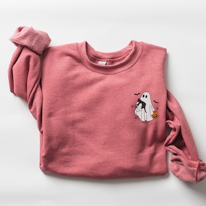 Op de afbeelding: Een roze sweatshirt met een witte geest die een zwarte kat en een pompoen vasthoudt. De geest wordt omringd door vleermuizen.