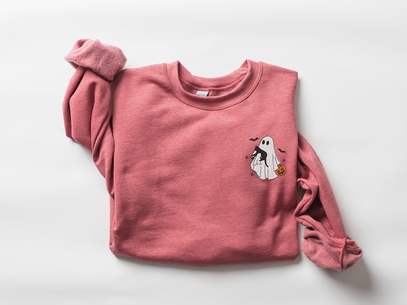 Op de afbeelding: Een roze sweatshirt met een witte geest die een zwarte kat en een pompoen vasthoudt. De geest wordt omringd door vleermuizen.