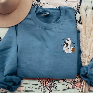 Op de afbeelding: Een blauw sweatshirt met een witte geest die een zwarte kat en een pompoen vasthoudt. De geest is geborduurd op de voorkant van het sweatshirt.