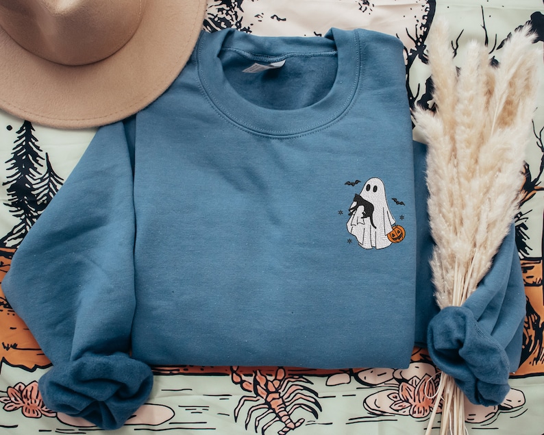 Op de afbeelding: Een blauw sweatshirt met een witte geest die een zwarte kat en een pompoen vasthoudt. De geest is geborduurd op de voorkant van het sweatshirt.