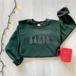 May include: A dark green sweatshirt with a white embroidered design of five Christmas trees. A red mug with the text "Merry & Bright" is in the bottom right corner of the image.