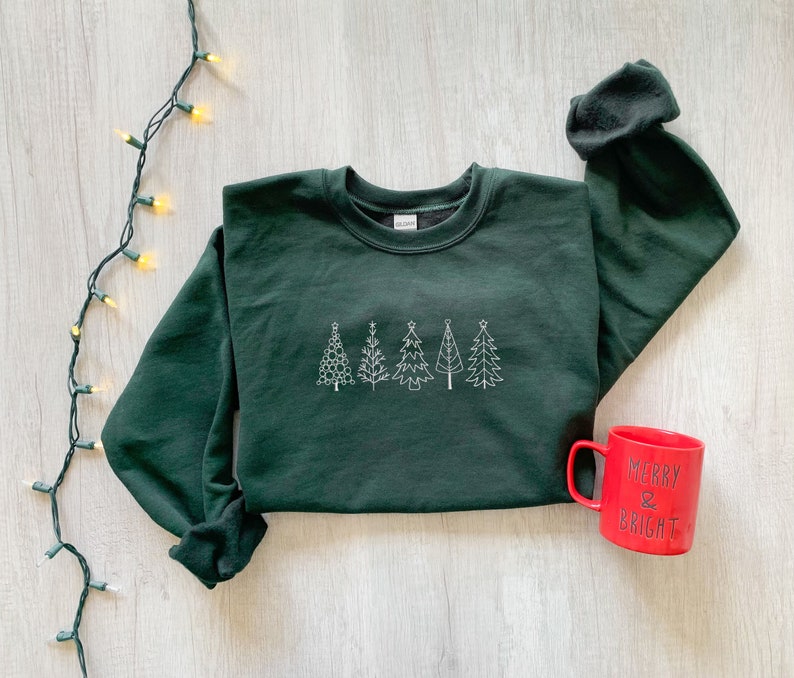 May include: A dark green sweatshirt with a white embroidered design of five Christmas trees. A red mug with the text "Merry & Bright" is in the bottom right corner of the image.