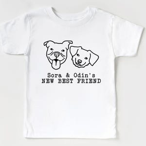 A white t-shirt with a graphic design featuring two dog faces and the text "Sora & Odin's NEW BEST FRIEND".