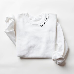 Op de afbeelding: Een witte crewneck sweatshirt met lange mouwen. Het woord "mama" is in een zwart script geschreven op de linkerbovenkant van de borst van de sweatshirt. De sweatshirt ligt plat op een wit oppervlak.