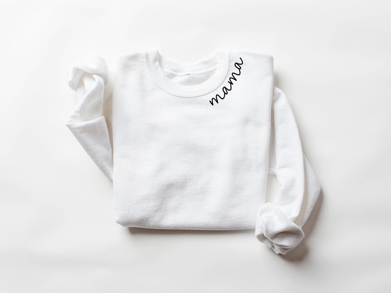 Op de afbeelding: Een witte crewneck sweatshirt met lange mouwen. Het woord "mama" is in een zwart script geschreven op de linkerbovenkant van de borst van de sweatshirt. De sweatshirt ligt plat op een wit oppervlak.