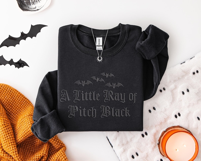 May include: Black crewneck sweatshirt with the phrase "A Little Ray of Pitch Black" and bat graphics. A silver necklace with a sun pendant is on the sweatshirt. Decorative bats and a skull are on the white surface. A candle and orange blanket are also visible.