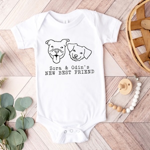 A white baby onesie with a graphic of two dogs and the text "Sora & Odin's NEW BEST FRIEND" printed on it, along with a wooden basket and some greenery.
