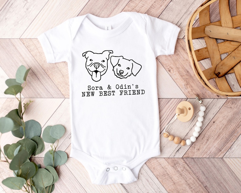 A white baby onesie with a graphic of two dogs and the text "Sora & Odin's NEW BEST FRIEND" printed on it, along with a wooden basket and some greenery.