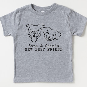 A gray t-shirt with a graphic design featuring two dog faces and the text "Sora & Odin's NEW BEST FRIEND".