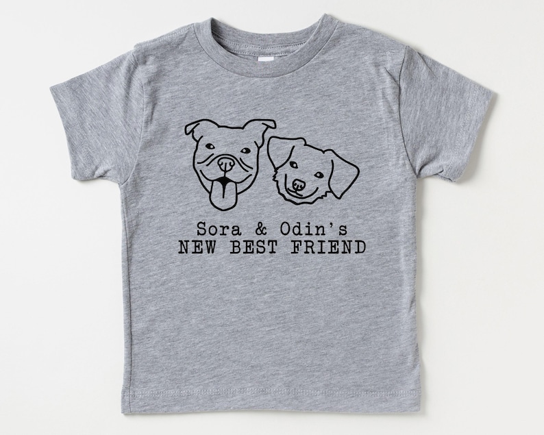 A gray t-shirt with a graphic design featuring two dog faces and the text "Sora & Odin's NEW BEST FRIEND".