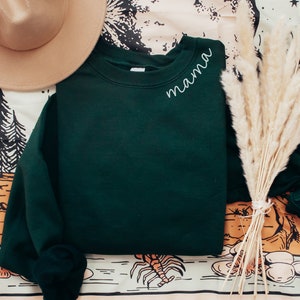 Op de afbeelding: Een donkergroene sweatshirt met het woord "mama" in witte letters op de linkerbovenborst. Een beige hoed en een bos gedroogd pampasgras liggen naast de sweatshirt. De achtergrond heeft een patroonstof met illustraties met een natuurthema.