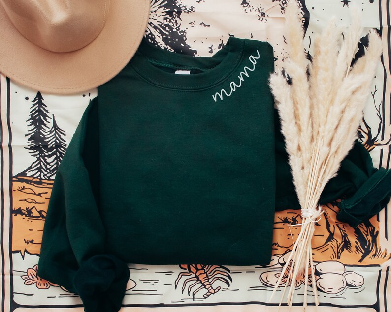 Op de afbeelding: Een donkergroene sweatshirt met het woord "mama" in witte letters op de linkerbovenborst. Een beige hoed en een bos gedroogd pampasgras liggen naast de sweatshirt. De achtergrond heeft een patroonstof met illustraties met een natuurthema.