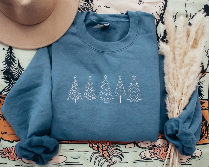May include: A blue sweatshirt with a white embroidered design of five stylised Christmas trees. The trees are arranged in a row across the front of the sweatshirt.