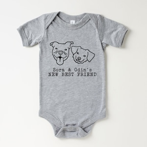A gray baby onesie with a graphic design of two dog faces and the text "Sora & Odin's NEW BEST FRIEND" printed on it.