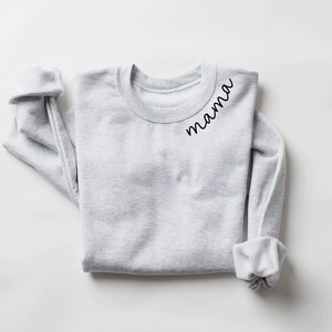 Op de afbeelding: Een lichtgrijs sweatshirt met het woord "mama" in zwart script gedrukt op de linkerbovenborst. De mouwen zijn opgerold. Het sweatshirt is netjes op een wit oppervlak gevouwen.