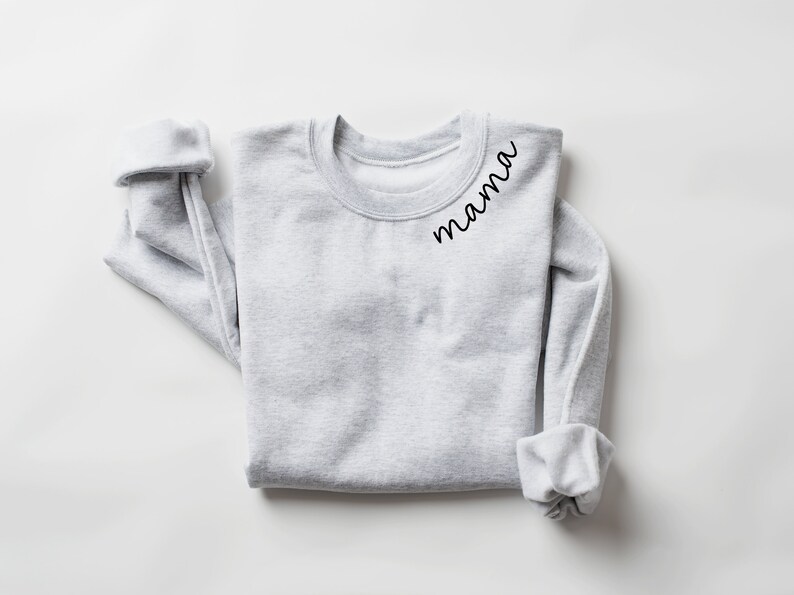Op de afbeelding: Een lichtgrijs sweatshirt met het woord "mama" in zwart script gedrukt op de linkerbovenborst. De mouwen zijn opgerold. Het sweatshirt is netjes op een wit oppervlak gevouwen.
