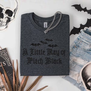 May include: Dark gray crew neck t-shirt with the phrase "A Little Ray of Pitch Black" and bat graphics. A silver chain necklace rests on the shirt. The shirt is paired with denim shorts and Halloween-themed decorations, including a skull and bats.