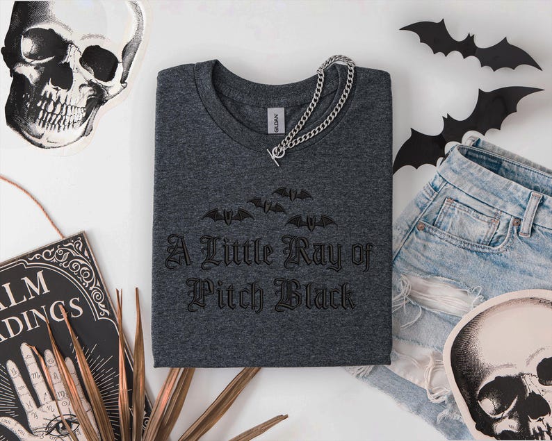 May include: Dark gray crew neck t-shirt with the phrase "A Little Ray of Pitch Black" and bat graphics. A silver chain necklace rests on the shirt. The shirt is paired with denim shorts and Halloween-themed decorations, including a skull and bats.