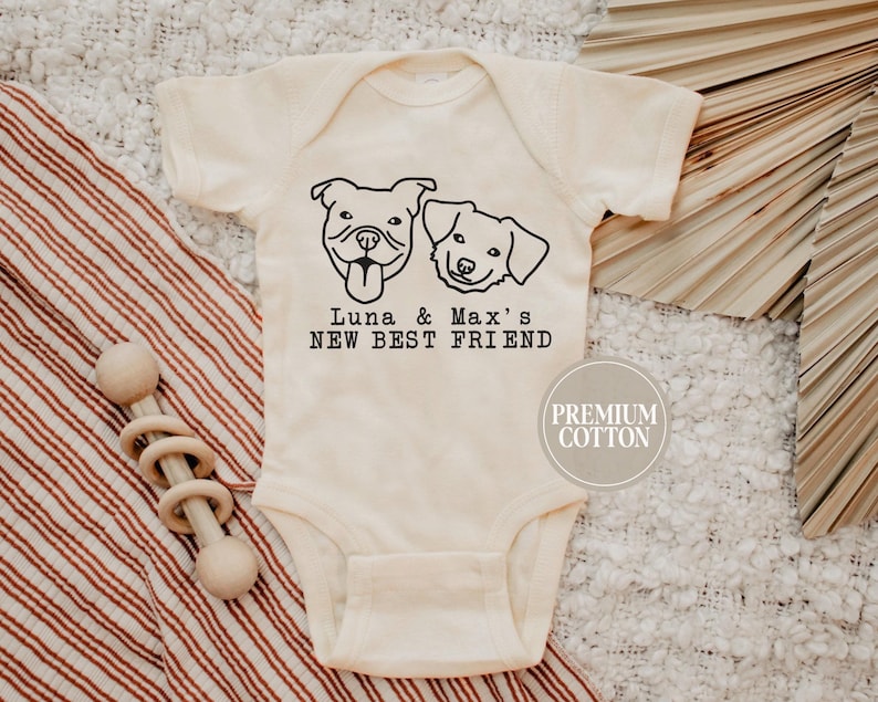 A baby onesie with a graphic of two dogs and the text "Luna & Max's NEW BEST FRIEND" printed on it, along with a "PREMIUM COTTON" logo.