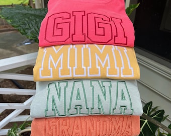May include: A stack of four crewneck sweatshirts in coral, yellow, light green, and orange. Each sweatshirt features embroidered block lettering in a darker shade of the fabric color, reading GIGI, MIMI, NANA, and GRANDMA respectively.