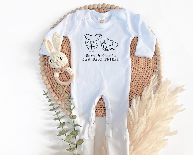 A white onesie with a graphic of two dogs and the text "Sora & Odin's NEW BEST FRIEND" is displayed on a woven basket, accompanied by a stuffed rabbit and some greenery.