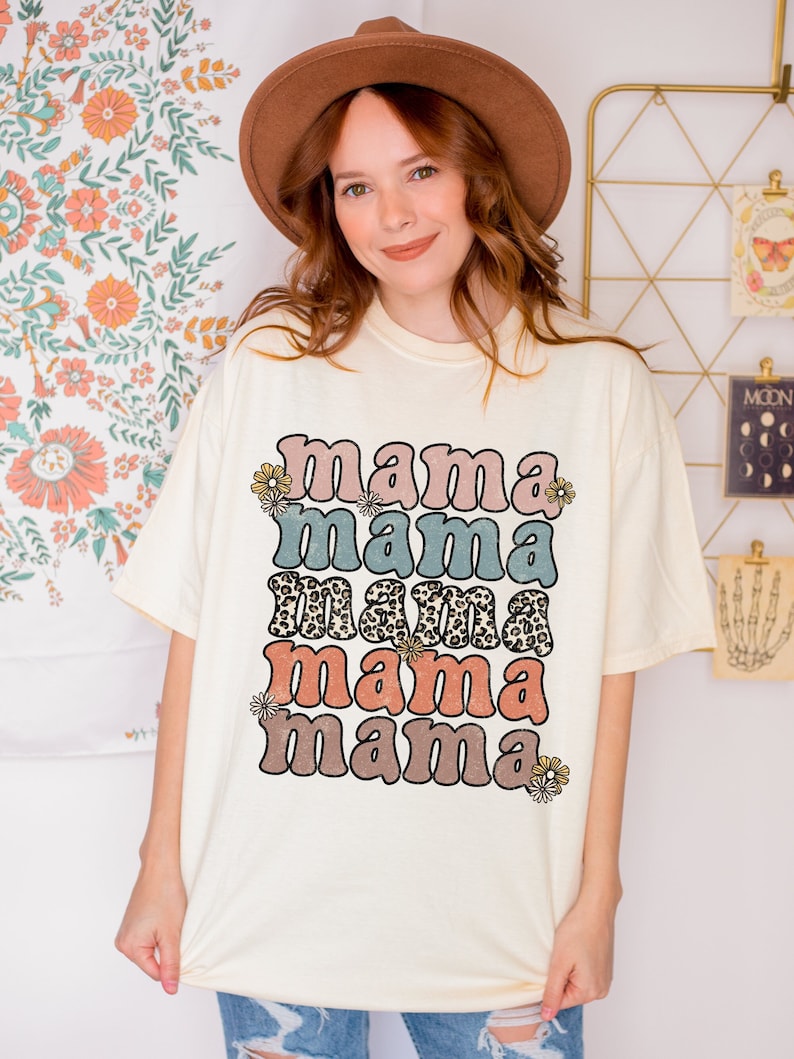 May include: A white t-shirt with the text "mama mama mama mama" in a retro font with different colors and patterns. The text is stacked vertically with a floral design on either side.