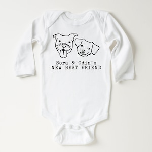 A white baby onesie with a graphic design of two dog faces and the text "Sora & Odin's NEW BEST FRIEND" printed on it.